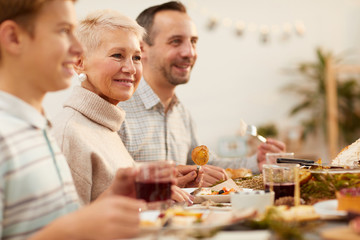 Mature happy woman sitting at the table between the members of her family and smiling while have family dinner at home