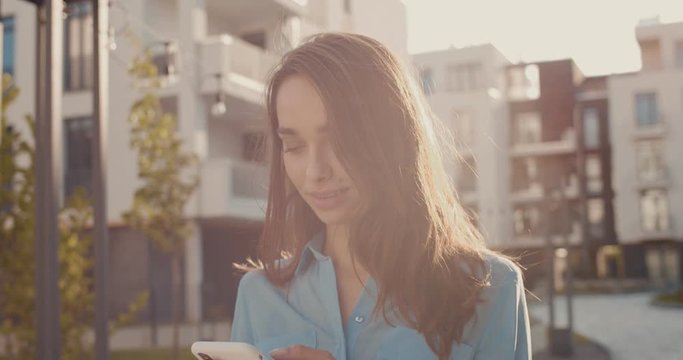 Close up of the Caucasian young pretty girl in sunlight and tapping or scrolling on the smartphone at the street among living block buildings. Outdoors.