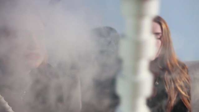 vacation. two young brunette girls with bright makeup in black clothes, sitting in cafe on gray couch and Smoking hookah. the girl lets out smoke nose, girls talking and smiling, slow motion, close up