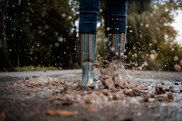 A woman wearing rubber boots jumps into a puddle on a cold autumn day.
