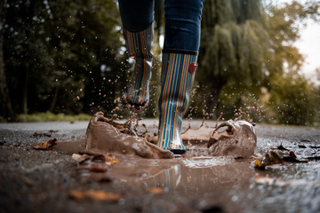 A woman wearing rubber boots jumps into a puddle on a cold autumn day.