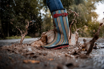 A woman wearing rubber boots jumps into a puddle on a cold autumn day.