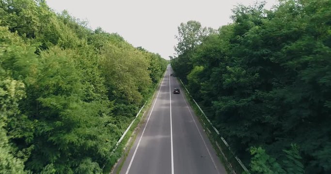 Aerial view: Cinematic shot. Flying over road with black car drives surrounded by forest. Car driving along the forest road. 4k