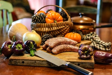 Selective focus still life of preparation of German bratwurst purple peppers and onions on wooden cutting board with a large knife, with blurred basket of pumpkins and pine cones in background 