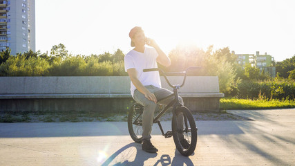 Young Caucasian male with cap talking on the phone and smiling on bmx bicycle in park on sunny summer day