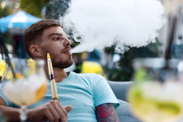 Handsome guy exhales hookah smoke. Young man smoking a fragrant oriental hookah outside. Rest with...