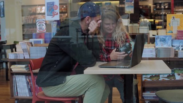 Shot through glass of young hipster generation z couple of blogger or social media influencers discussing content from professional camera in cosy cafe or coffee shop. Millennial lifestyle