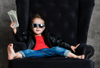 Impudent kid boy millionaire in leather jacket and red t-shirt is sitting lounging in armchair...