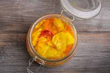 Homemade pickled cabbage leaves and pepper and carrots in a glass jar with an open lid on a wooden brown background. Rustic style