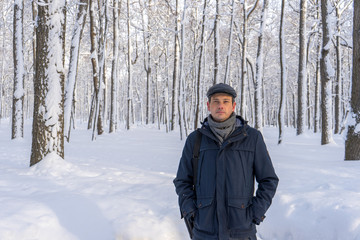 Fototapeta na wymiar Portrait of handsome middle-aged man walking in winter snowy park or forest. Attractive man in jacket, scarf and cap looking at camera. Winter mood, authentic lifestyle concept, stylish male outfit