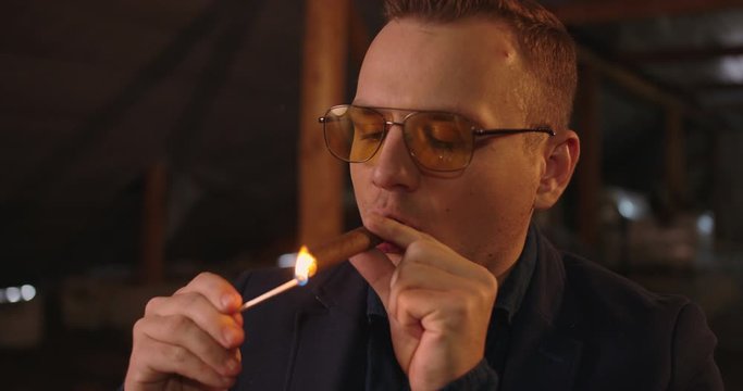 Portrait of handsome elegant man in glasses and blazer , He sets fire his cigare. Dark room location , 