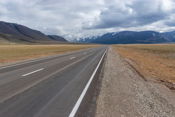 The gray asphalt road goes far and abuts against blue-blue clouds and mountains on a cloudy autumn day in the Altai Mountains