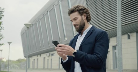 Attractive Caucasian Businessman standing near modern International airport. Young bearded Man texting Messages and smiling. Stylish Man using Smartphone in Airport. International Business. Men. App.
