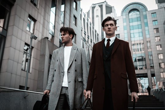 Two fashion men models posing on city street. Wearing in classic and casual closes. Suit, coat, shirt, sweater, boots and leather bag. Men model test near business center