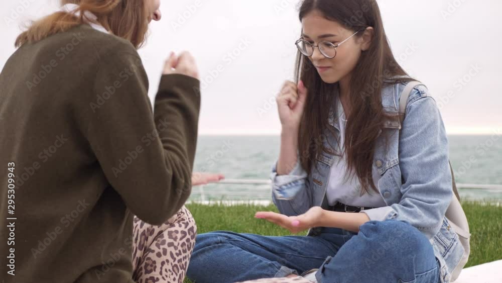 Sticker Cheerful laughing young girls friends playing rock-paper-scissors and having fun while sitting at the pier near the sea