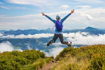 Girl with a backpack that makes a high jump for joy on the top of a high mountain above the clouds