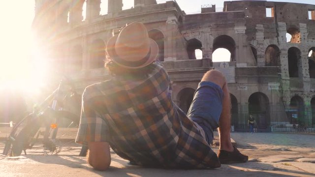 Happy young man tourist wearing shirt and hat with bike sitting and taking selfies with vintage camera at colosseum in Rome, Italy at sunrise.