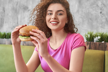 Hungry woman smiling while eating tasty hamburger in cafe