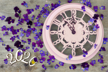 2020 numbers on a wooden background next to purple pansies flowers and a pink clock showing five minutes to midnight