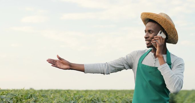 Portrait of the happy young African American man in a hat and apron standing in the field in summer and speaking cheerfully on the mobile phone.