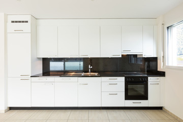 Empty room with white walls, travertine floor and white kitchen and black marble