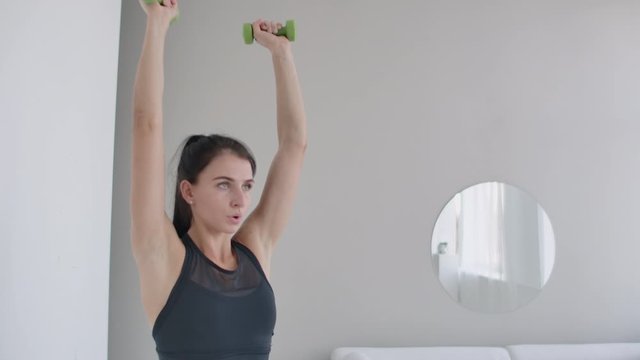A young Caucasian woman lifts dumbbells for arm and shoulder exercises. Lift dumbbells over your head