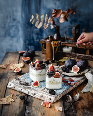 jars with creamy yogurt, oats, figs and berries topped with honey on blue wooden board with fresh figs on plate, textile, breakfast concept