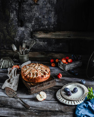 apple and plum pie on wooden board among fresh fruits, kitchen utensils on grey wooden table