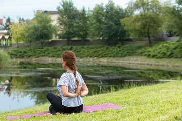 Young beautiful woman doing yoga exercise in green park near the pond. Exercices for improve the flexibility. Wellbeing and wellness. Healthy lifestyle and fitness concept.