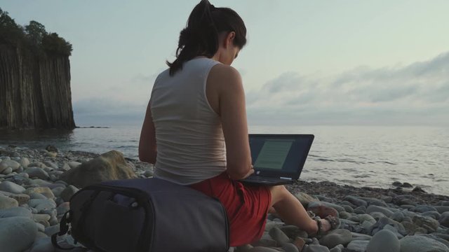 A woman is sitting on the beach at sunset with a laptop and typing. Woman on the beach working on a laptop rocks and sea in the background.