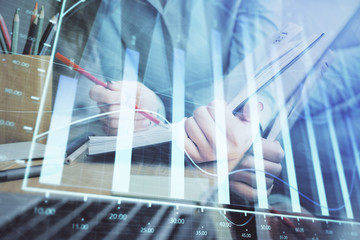 A woman hands writing information about stock market in notepad. Forex chart holograms in front. Concept of research. Multi exposure