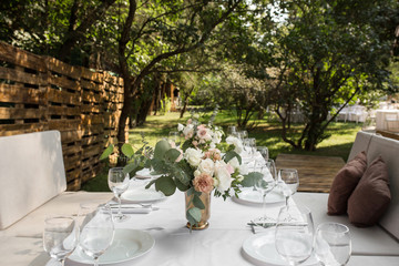 Wedding table setting decorated with fresh flowers in a brass vase. Wedding floristry. Banquet table for guests outdoors with a view of green nature. Bouquet with roses, eustoma and eucalyptus leaves
