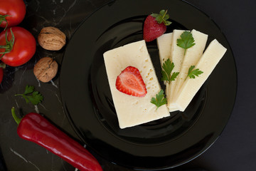 White cheese on a black plate, decorated with strawberries, red pepper, dill, cherry tomatoes and walnuts, on a dark background. The concept of cheese products.