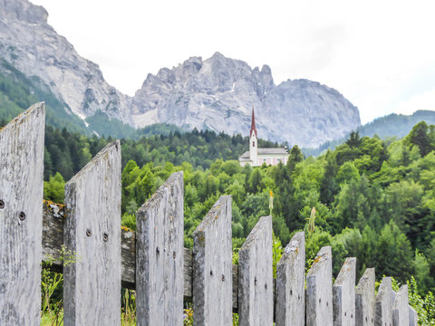 A church popping out of the forest, with a tall, sharp and rocky mountains behind. The church is build on a rock, making it taller than surrounding trees. There is a fence in front.