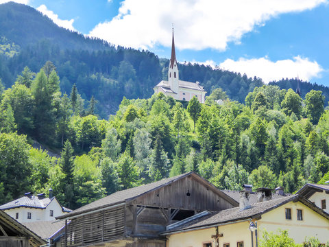 A church popping out of the forest, with a tall, sharp and rocky mountains behind. The church is build on a rock, making it taller than surrounding trees. There is a small village in the valley.
