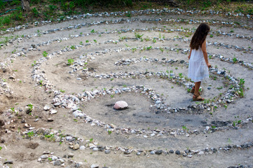 A young woman with long brown hair and white dress is walking away from the center of a maze or stone labyrinth.