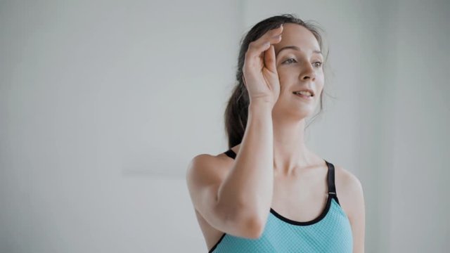 Tired pretty young brunette caucasian girl with pigtail wearing blue fitness top wipes sweat from her forehead after effective workout on white background, smiles and laughs. Healthy lifestyle
