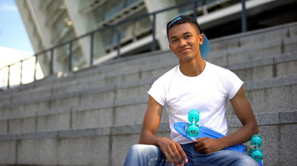 Mixed-race teenager with skateboard smiling into camera, leisure activity, hobby