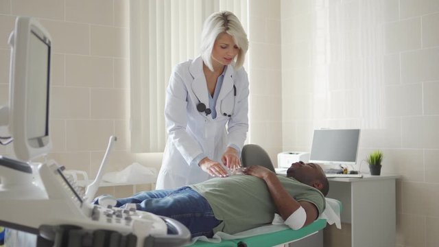 Tracking shot of female physician in white coat palpating belly of young African patient with bandage on arm after venipuncture lying on couch in doctors office