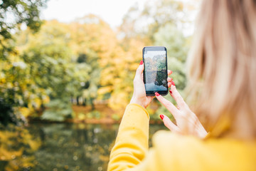 Blonde woman it yellow coat makes photo on cell phone in the park in autumn.Close up, selective focus.