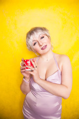 pretty curvy young blonde woman with short hair enjoys drinking tea from cute red polka dot ceramic cup on yellow studio background