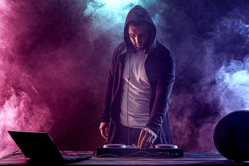 Young stylish man in glasses posing behind mixing console on colored smoke studio background.