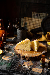 homemade two tasty slices of baked lemon biscuit cake with powdered sugar on top stands on wooden board on rustic table with lemons, old bottles opposite concrete wall, selective focus