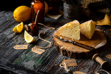 homemade two tasty slices of baked lemon biscuit cake with powdered sugar on top stands on wooden board on rustic table with lemons, old bottles opposite concrete wall, selective focus