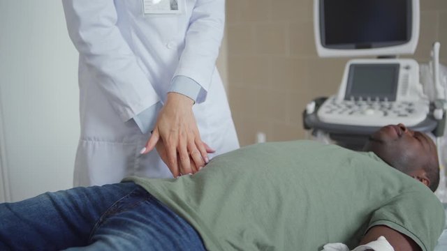 Tilt up and down shot of female doctor in white coat palpating stomach of African patient lying on couch and suffering from pain