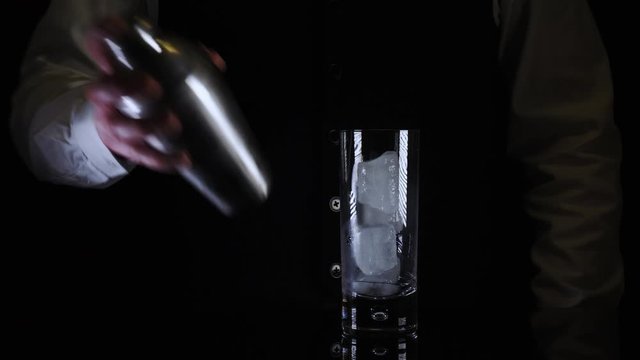 Bartender mixes a Bloody Mary drink cocktail in a shaker and pours into a glass with ice cubes and a celery against a black background
