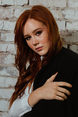 Portrait of a girl with red hair near a white brick wall. In a light blouse and black trousers. Business style clothing