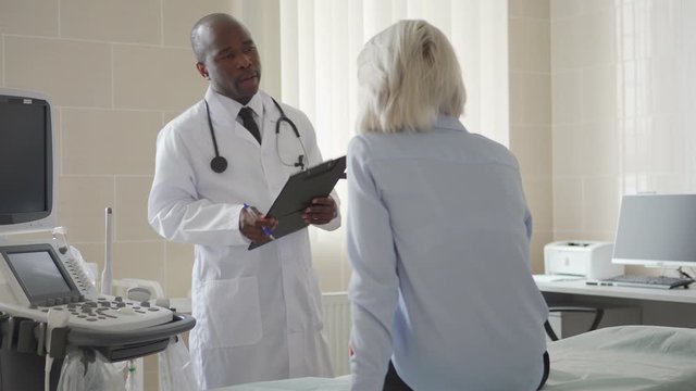 Back view of blonde female patient sitting on couch in doctors office and listening to African American physician in white coat holding clipboard and explaining diagnosis to woman