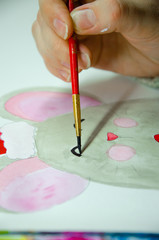 Girl sitting draws with watercolors on a white sheet of paper a mouse with a green gift. Top view. New Year 2020, the year of the mouse