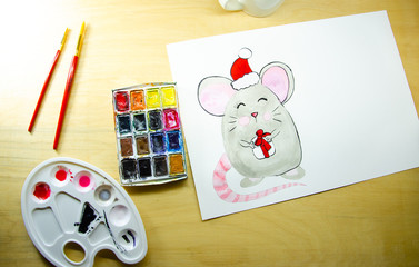 Girl sitting draws with watercolors on a white sheet of paper a mouse with a green gift. Top view. New Year 2020, the year of the mouse